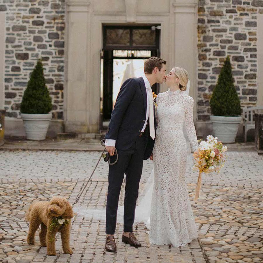 Groom in navy suit and holding dog on leash kisses bride in a long-sleeve lace gown holding a bouquet